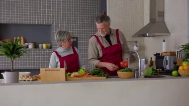 Mutlu emekli çift önlük giyip modern mutfakta taze sebzelerle sağlıklı bir yemek hazırlıyor, sohbet ediyor ve birlikte yemek pişiriyorlar.