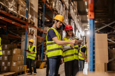 Güvenlik yelekleri ve başlık takan depo çalışanları barkod tarayıcıları ve dijital tabletlerle paketleri tarıyor, yoğun bir dağıtım merkezinde envanter ve lojistiği yönetiyorlar.