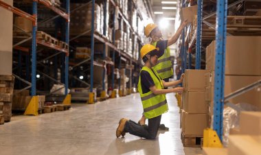 Güvenlik yelekleri ve kask giyen iki depo işçisi geniş bir dağıtım deposunun koridorlarında gezinirken aktif olarak stok ve stok seviyelerini kontrol ediyorlar.