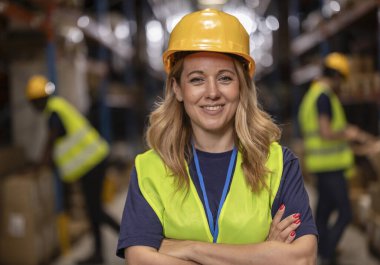 Kollarını kavuşturup gülümseyen, güvenlik yeleği ve şapka takan, büyük bir depoda lojistik ve dağıtım yöneten, arka planda çalışan meslektaşları olan kendinden emin kadın yönetici.