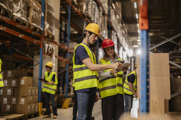 Работники склада в защитных жилетах и шляпах сканируют пакеты со сканерами штрих-кода и цифровыми планшетами, организуют инвентаризацию и логистику в оживленном центре дистрибуции.