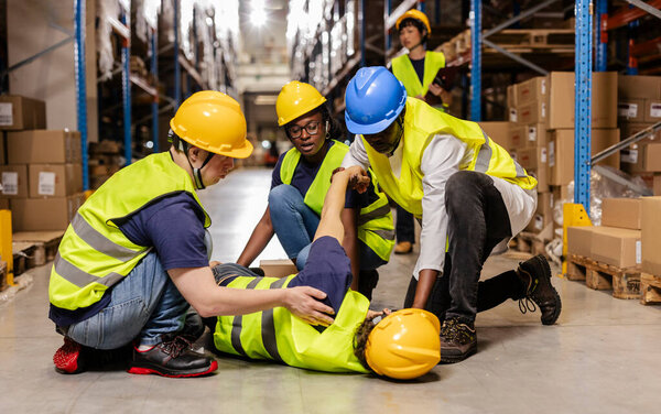 Warehouse workers assisting injured colleague lying on floor after workplace accident, providing first aid and support in distribution center