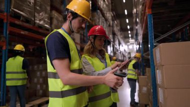 Güvenlik yelekleri ve kask giyen depo çalışanları, büyük dağıtım merkezi içinde verimli bir envanter yönetimi sağlamak için barkod tarayıcıları ve dijital tabletler kullanarak paketleri tarıyorlar.