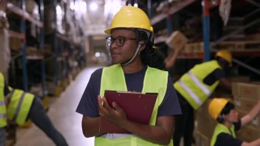 Güvenlik kaskı ve yeleği takan kadın denetçi, hareketli bir depodaki operasyonları dikkatle izliyor, arkaplandaki paketlerle işçiler ilgilenirken pano elinde.