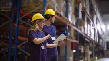 Koruyucu miğfer ve kimlik kartı takan iki depo çalışanı, istiflenmiş raflar arasında dijital tablete danışarak, muhtemelen envanter ve işlem emirlerini yöneterek,