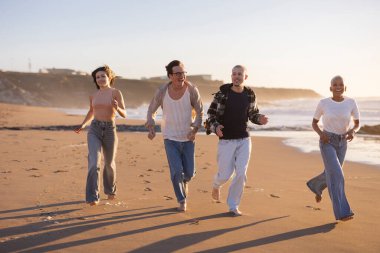 Dört neşeli arkadaş kumlu sahil boyunca neşeli bir koşu yapıyorlar, batan güneşin sıcak altın ışığında banyo yapıyorlar, neşeli ve kaygısız bir atmosfer yaratıyorlar.