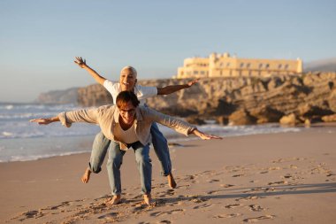 Adam kumsalda bir kadını sırtında taşıyor. İkisi de kollarını uçak kanadı gibi şaklabanca açıyor. Deniz kenarında romantik bir anın tadını çıkarıyor.