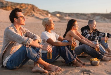 Dört arkadaş kumlu sahilde oturmuş, birlikte gün batımını izlerken içkilerin ve abur cuburların tadını çıkarıyorlar. Kaygısız yaz akşamlarının ve neşe dolu birlikteliğin özünü somutlaştırıyorlar.