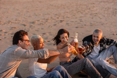 Bir grup neşeli çoklu etnik arkadaş plajda rahatlatıcı bir akşamın tadını çıkarıyor, güneş batarken bira şişeleriyle kadeh kaldırıyor, sıcak ve şenlikli bir atmosfer yaratıyorlar.