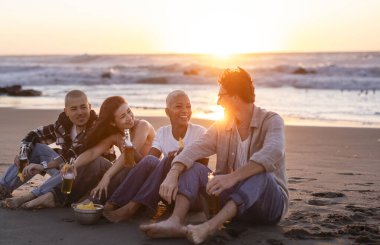 Dört arkadaş gün batımında kumsalda oturmuş, içeceklerin, abur cuburların ve arkadaşlığın tadını çıkarıyor, sıcak ve neşeli bir atmosfer yaratıyorlar.