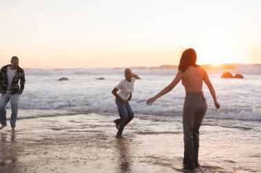 Üç arkadaş okyanus dalgalarında koşuyor ve oynuyorlar, kumlu bir sahilde güzel günbatımının tadını çıkarıyorlar, tasasız ve neşeli bir atmosfer yaratıyorlar.
