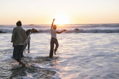 Bir grup arkadaş kıyı şeridi boyunca yürür, okyanus dalgalarında su sıçratır altın saat boyunca baş döndürücü bir güneşin altında güneşlenirken mutluluk ve kaygısızca neşe saçar.