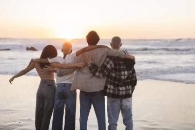 Birbirlerine sarılan dört genç arkadaş kumsalda güzel bir gün batımının tadını çıkarıyorlar. Birbirlerine yakınlık ve sevgi dolu bir ortam yaratıyorlar.