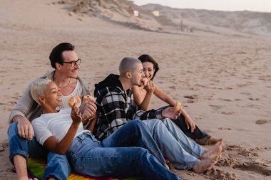Dört arkadaş plajda canlı bir battaniyeye oturmuş, nefis çöreklerin tadını çıkarırken, gün batımının tadını çıkarıyorlar, neşe ve beraberliği somutlaştırıyorlar.