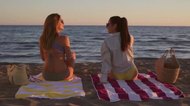Renkli plaj havlularında dinlenen iki bayan arkadaş, altın saat boyunca sakin okyanus sularının yanında huzurlu anların tadını çıkarırken, gün batımının ışıl ışıl renklerine bürünüyorlar.