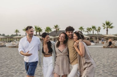 Gün batımında kumsalda yürüyen beş neşeli arkadaş, birlikte tatilin tadını çıkarırken gülüyorlar ve arkadaşlığın sıcaklığını kucaklıyorlar.