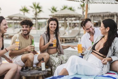 Bir grup neşeli arkadaş gülüyor, içiyor ve plaj otelinde birlikte dinleniyorlar. Yaz tatillerinin tadını çıkarıyorlar. Plaj şemsiyesi altında.