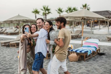 Beş neşeli arkadaş kumlu bir plajda gün batımının tadını çıkarıyorlar, bira şişeleri tutarken ve tatillerini birlikte kutlarken neşeli bir selfie çekiyorlar. Kahkahalar ve gülümsemeler havayı dolduruyor