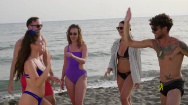 Mayo giyen arkadaşlar beşlik çakıyor, gülüyor ve kaynaşıyor güneşli sahilde neşeli anları paylaşıyorlar, arkadaşlığı ve tatil mutluluğunu kutluyorlar.