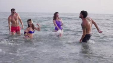 Renkli mayo giyen genç arkadaşlar yaz tatilinde parlak okyanus dalgalarında yüzerken birlikte oynayıp, sıçrayıp, kaygısız anların tadını çıkarıyorlar.