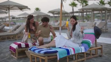 Üç arkadaş, iki kadın ve bir erkek plaj sandalyelerinde dinleniyor, içki içiyor, sohbet ediyor ve güneşli bir kumsal tatilinde birlikte gülüyorlar.