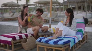 Plaj sandalyelerinde rahat bir şekilde uzanan arkadaşlar, tropik deniz kıyısındaki altın günbatımı anlarında koruyucu güneş şemsiyesi altında kahkahalar ve şişe içecekleri paylaşıyorlar.