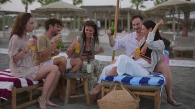 Mutlu turistler plajda rahatlatıcı bir akşamın tadını çıkarıyorlar, plaj sandalyelerinde oturup şişeler ve bardaklarla kadeh kaldırıyorlar, kahkahalar atıyorlar ve güzel zamanlar geçiriyorlar.
