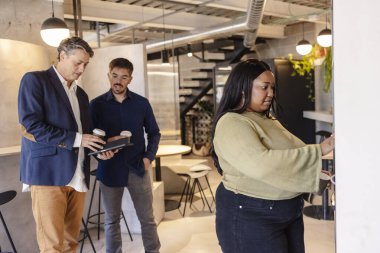 Modern ofis ortamında bir mola sırasında dijital tablet kullanan ve kahve içen çok ırklı iş adamları bir proje üzerinde işbirliği yapıyorlar.