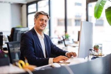 Modern bir ofis masasında oturmuş, şık bir takım elbise giyerken bilgisayar kullanan gülümseyen işadamı, pencereden parlak şehir manzaralı kameraya güvenle bakıyor.