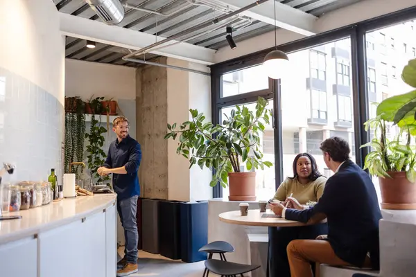 İş ortakları modern ofis dinlenme odasında strateji tartışırken, bir meslektaşımız arka planda bulaşıkları yıkıyor, takım çalışmasını ve rahat bir çalışma ortamını ön plana çıkarıyor.