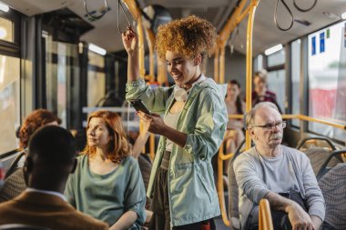 Boynunda kıvırcık saçları ve kulaklıkları olan genç bir kadın kalabalık bir otobüste akıllı telefonunu denge için korkuluklara tutunurken gülümsüyor.