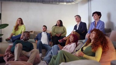 Çok kültürlü bir grup arkadaş, birlikte film izlerken keyifli anları paylaşıyor, sıcak atmosferi ve gerçek bağlantıları olan sıcak oturma odasında gülüyor ve alkışlıyorlar.