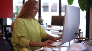 Masada çalışan, yeşil kazaklı, odaklanmış kızıl saçlı iş kadını. Parlak, modern ofis çalışma alanının içindeki bilgisayar klavyesine yazı yazıyor.