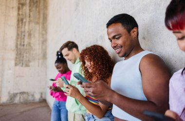 Bir grup farklı arkadaş, beton duvara yaslanırken akıllı telefonları kullanıyor, sosyal medyanın ve gelişigüzel bir şehir ortamında online iletişimin tadını çıkarıyor.