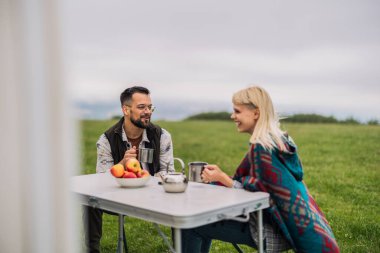 Genç çift rahat bir kamp deneyiminin tadını çıkarıyor, kırsal bir masada oturuyor, metal bardaklardan sıcak içecekler yudumluyor, huzurlu ve bulutlu bir günde, kahkahalarını ve taze elmalarını paylaşıyor.