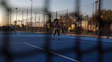 Genç bayan sporcu, mavi tenis kortunda servis alıyor. Güneşli bir öğleden sonra ağdan görülüyor. Yeteneklerini çalışıyor ve spordan zevk alıyor.
