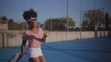 Pembe tişörtlü, beyaz şortlu profesyonel siyahi kadın tenisçiyi yakalamak, mavi tenis kortunda güçlü bir servis hazırlamak, atletik beceri ve konsantrasyon göstergesi