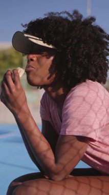 Saha kenarında oturan Afro-Amerikan sporcu kadın, hızlı protein barının keyfini çıkarıyor, güneş ışığı, huzurlu bir öğleden sonra tatilinde çitlerden dramatik gölgeler düşürüyor.