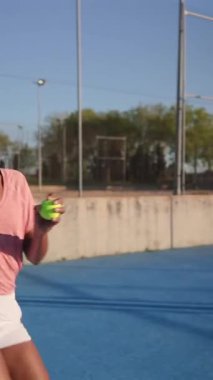 Kendine güvenen zenci kadın tenisçi maç boyunca hizmet etmeye hazırlanıyor, güçlü atletik duruş sergiliyor ve güneşli tenis kortuna yoğun bir şekilde odaklanıyor