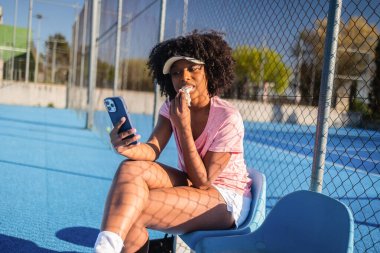 Afrikalı Amerikalı tenis oyuncusu sahada mola veriyor, abur cubur yiyor ve mavi plastik bir sandalyede otururken akıllı telefonunu karıştırıyor.
