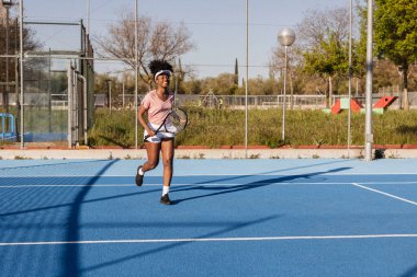Genç siyahi bir kadın tenis maçında elinde raketiyle koşuyor ve gülümsüyor. Arka planda ağ ve çitle.