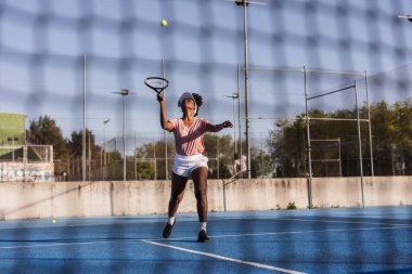 Profesyonel bayan tenisçi parlak güneşli bir gökyüzü altında canlı mavi sert bir kortta topa servis yapıyor. Rekabetçi maçı kazanmak için odaklanma ve kararlılık gösteriyor.