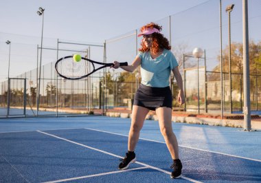 Kızıl tenli tenisçi, canlı bir mavi kortta raketiyle topa vuruyor. Spor, yarışma ve maçın heyecanıyla dolu güneşli bir günün tadını çıkarıyor.