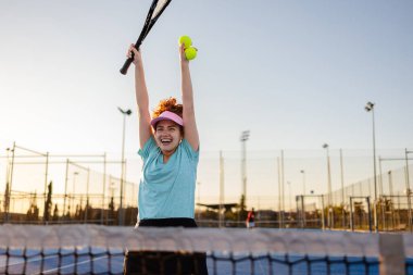 Kızıl saçlı tenisçi zaferi yükseltilmiş raket ve toplarla kutluyor. Güneşli tenis kortunda neşe ve heyecan saçıyor. Rekabet ve başarı ruhunu somutlaştırıyor.