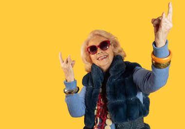 Kırmızı güneş gözlüğü takan ve şık giyinen yaşlı bir kadın coşkuyla rock 'n roll tabelaları yapıyor, enerji yayıyor ve hayat dolu sarı bir arka plana karşı sonsuz serinlik sergiliyor.