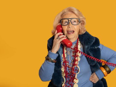 Gözlükleri ve şık aksesuarları olan zarif son sınıf öğrencisi bir kadın, canlı sarı bir arka plana neşe ve incelik saçan bir telefon görüşmesiyle mutlu mesut meşgul.