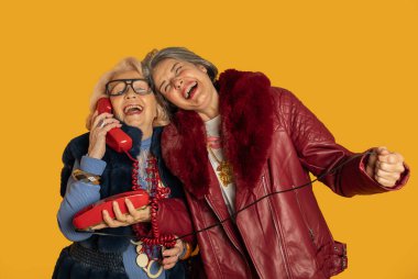 İki modaya uygun son sınıf öğrencisi telefonda neşeli bir konuşma yapıyorlar. Canlı sarı bir fon karşısında gülüp eğleniyorlar.