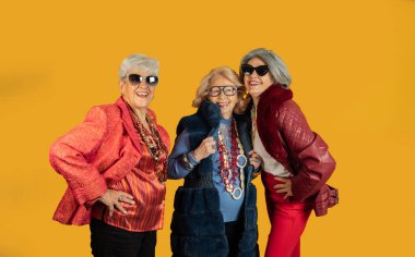 Renkli kıyafetler, güneş gözlükleri ve kolyeler takan üç moda son sınıf öğrencisi, canlı sarı bir arka planda poz veriyor ve gülümsüyor, güven ve neşe saçıyor.