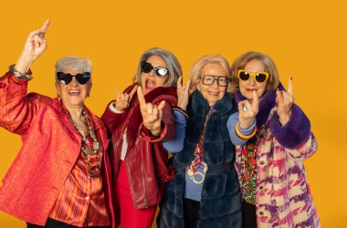 Renkli kıyafetler ve güneş gözlüğü takan dört moda son sınıf öğrencisi, parlak sarı arka planda birlikte eğlenirken rock 'n roll tabelaları yapıyor.