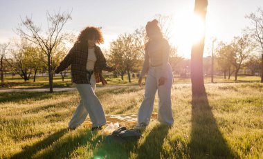 İki mutlu genç kadın gün batımında parkta dans ediyor, çim ve ağaçlarla çevrili bir battaniye ve sırt çantasıyla pikniğin tadını çıkarıyorlar, arkadaşlıkları ve boş zamanları kutluyorlar.